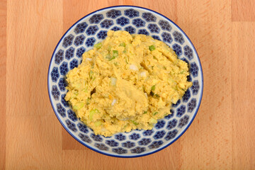 A top-down close-up shows a blue and white patterned bowl filled with a creamy, light yellow mixture of mashed yolks, avocado, and chopped onions, prepared for deviled eggs, on a wooden surface