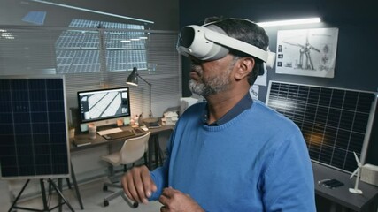 Chest up shot of senior male engineer wearing augmented reality headset and enhancing solar power system at site at renewable energy office with technical drawing, solar panels and computer in back - Powered by Adobe