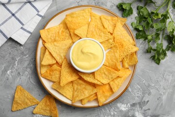 Tasty cheese dipping sauce in bowl, nacho chips and parsley on grey table, flat lay