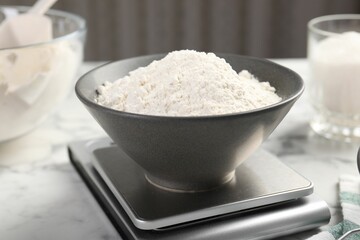 Digital kitchen scale with bowl of flour and other products on white marble table, closeup