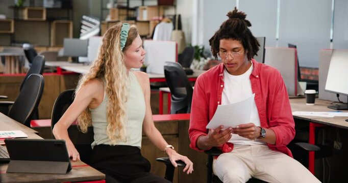 Diverse coworkers reviewing printed documents in modern office, with monitors and coffee cups