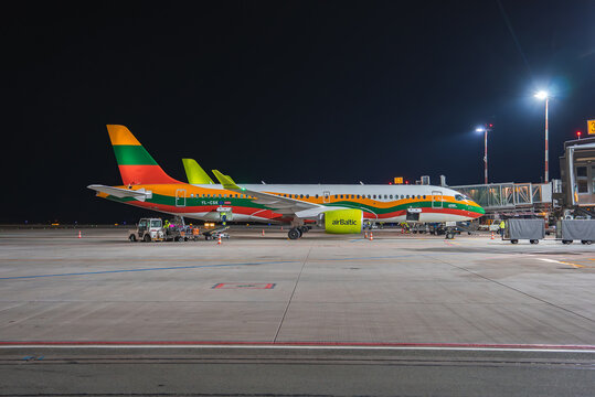 An airBaltic airplane with green, yellow, and red stripes sits on an airport tarmac at night. Ground vehicles and a jet bridge are visible under bright lights.
