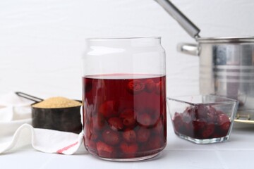 Tasty cherry compot in glass jar and ingredients on white tiled table, closeup