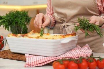 Woman putting rosemary onto tasty lasagna at white table indoors, closeup