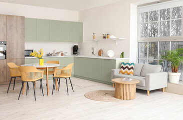 Interior of light spacious kitchen studio with dining table, armchairs, mimosa flowers, houseplant, counters and sofa