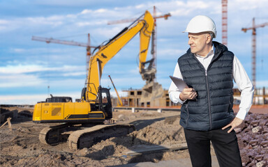 Construction manager with tablet inspects building site with excavator and cranes in background, supervising progress and heavy equipment work – Construction. Machinery repair. - obrazy, fototapety, plakaty