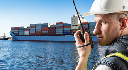 Port worker in helmet uses walkie-talkie while monitoring cargo ship arrival loaded with...
