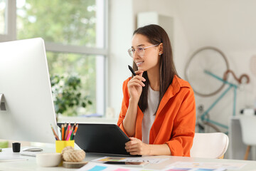 Thoughtful young female graphic designer with tablet sitting at table in office