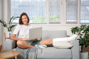 Young woman with broken leg using laptop on sofa at home