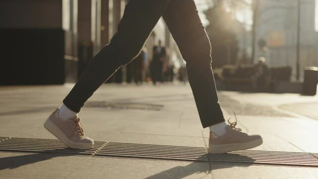 Side view gimbal following female feet steps walking pedestrian crossing street, sunny day, young woman wearing black pants and sneakers goes crosswalk. Low angle people legs stepping zebra asphalt