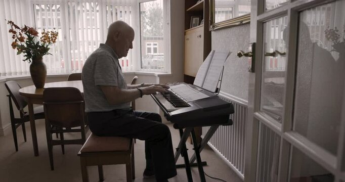 Senior man playing music on a keyboard with sheets in living room for entertainment in his house. Music, hobby and elderly guy in retirement with a song book to play on the piano in lounge at home.