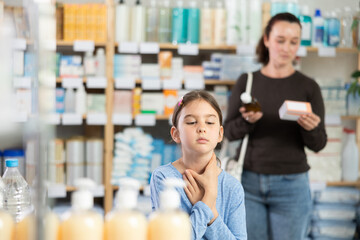 Child stands in the pharmacy with a sore throat and cough, and the mother chooses medicines and...
