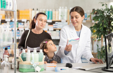 Obraz premium Smiling friendly young female pharmacist in white coat assisting woman, visiting pharmacy with preteen daughter, selecting medications and checking availability through computer system
