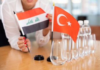 Little flag of Turkey on table with bottles of water and flag of Iraq put next to it by positive young woman in meeting room