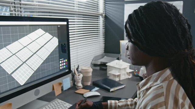Arc shot of young female African American solar engineer designing digital solar panel system model on computer using drawing tablet at desk, working in modern renewable energy office
