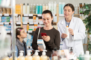 Woman and girl daughter in pharmacy scans QR code on cream and uses link to go to manufacturers website. Bored child pulls mother by hand, looks at goods. Pharmacist offer analogue remedy
