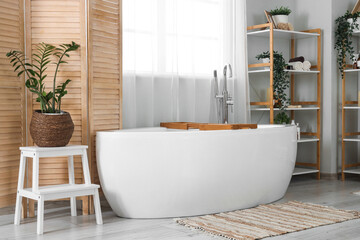 Interior of light bathroom with bathtub, wooden folding screen and shelf units
