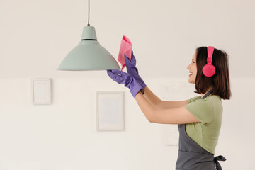 Female janitor in headphones cleaning hanging lamp at home