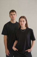 Obraz premium A minimalist studio portrait of two young people standing against a plain off-white wall. Both are wearing plain black t-shirts and dark pants. 