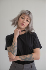 A portrait photograph of a person with silver-gray wavy shoulder-length hair against a plain light gray background. They are wearing a plain black crew neck t-shirt and gray pants.