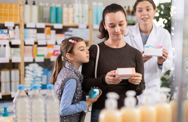 Interested woman with small daughter thoughtfully examining box of medicine while female pharmacist politely offering assistance in background in modern pharmacy