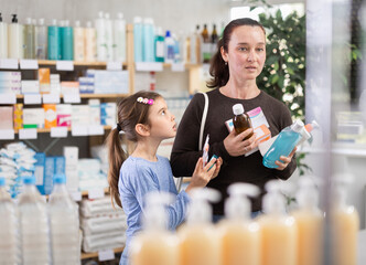 Woman stands in a pharmacy with daughter and holds various goods in hands. Customer is provided with a mouthwash, medicinal syrup and medical cream against the background of the shelves in the