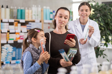 Interested woman with preteen daughter scanning packages of toothbrushes using phone, checking details online and comparing options while friendly female pharmacist explaining features in drugstore