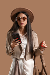 Fototapeta premium Stylish young woman in sunglasses using mobile phone on brown background