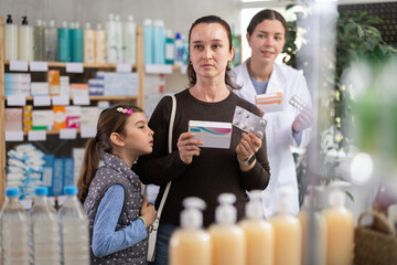 Focused woman with preteen daughter selecting medication at local pharmacy while friendly female pharmacist in background offering product options