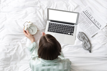 Woman in pajamas with alarm clock and blank laptop on bed, top view