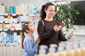 Woman stands with daughter in a pharmacy and makes purchases using an online application. Customer with a child scans the barcode of a plastic shampoo bottle to read the instructions on the website