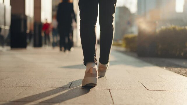 Close-up back view gimbal following shot female feet steps walking busy sidewalk, sunny day, young woman wearing black pants and white sneakers goes city street. Low angle people legs stepping avenue