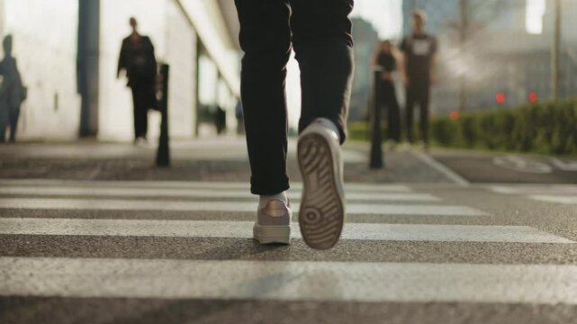 Back view following shot female feet steps walking pedestrian crossing street, sunny day, young woman wearing black pants and sneakers goes crosswalk. Low angle people legs stepping zebra asphalt