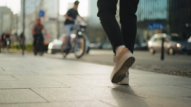 Close-up back view gimbal following shot female feet steps walking sidewalk, sunny day, young woman wearing black pants and white sneakers goes city street. Low angle people legs stepping avenue