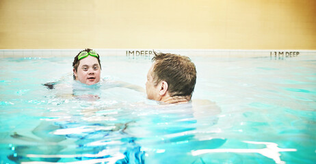 Man with down syndrome swimming in a pool during a sports class with a swim instructor. Disabled adult with a happy, healthy and active lifestyle doing a water training exercise with eye goggles.