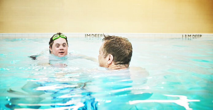 Man with down syndrome swimming in a pool during a sports class with a swim instructor. Disabled adult with a happy, healthy and active lifestyle doing a water training exercise with eye goggles.