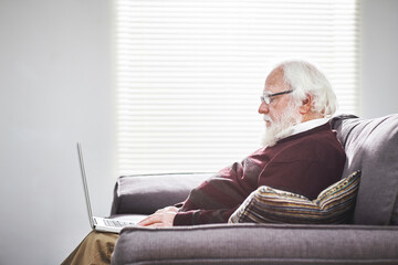 Relaxed senior man typing on a laptop relaxing on a couch at home and learning to use technology. A...
