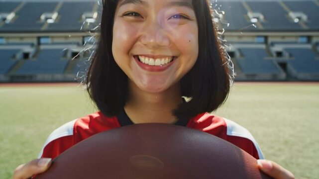 Young happy teenage asian girl wearing a red and white sports jersey holding an american football, rugby player smiling and looking at camera while standing in empty stadium on a sunny day. Dolly in