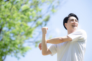 スポーツの準備運動や腕やストレッチを新緑と青空の中する男性
