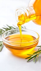 Golden oil pours from a glass bottle into a clear bowl, beside sprigs of rosemary