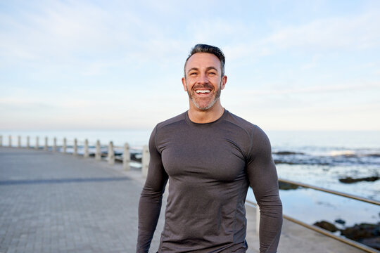 Mature man, runner and smile on promenade by ocean, break or pride with portrait for workout in morning. Person, training and fitness by sea with confidence, happy or outdoor for exercise in Spain