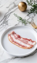A single crispy slice of bacon rests on a plain white plate atop a marble table. Gold decorative ornaments and greenery add a touch of elegance to the setting