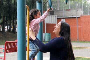 Obraz premium Divorced single mother plays with her 4-year-old Latina daughter, brunette with curly hair, on park equipment like the monkey bars to celebrate Mother's Day