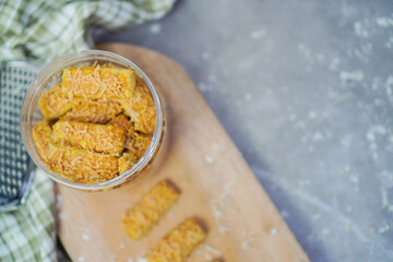 top view of an open jar filled with cheese cookies and some cookies lined up outside the jar, with cheese grater, gingham fabric and wooden chopping board below it