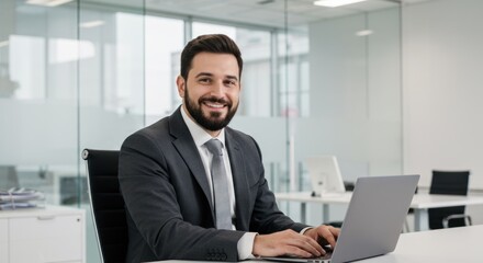 Man in suit sits at desk using laptop in modern office.