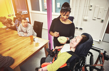 Nurse helping a disabled woman eat food at a healthcare home or institute for disability. Caregiver or volunteer working with a special needs girl in a wheelchair at a nursing clinic and having lunch