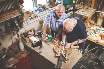 Senior craftsmen making a custom guitar in a workshop together helping and advising. Skilled mature or old carpenters working and building a string instrument in a carpenter shop