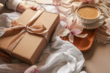 Woman holding a gift box with a satin ribbon on a cozy spring setup featuring magnolia flowers, a cup of cappuccino, and soft fabrics