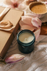 A minimalistic soy candle in a dark glass jar next to a kraft gift box with a satin ribbon and soft magnolia flowers, styled on white embroidered fabric in warm natural light