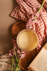 Top view of an unlit beige candle with pink hyacinths, open vintage book, wooden tray, soft fabric, and gingham cloth in warm, cozy spring atmosphere